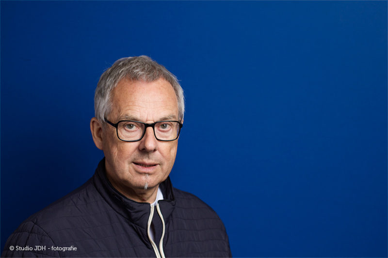 (Zakelijke) Portretfoto met kleur | Man met bril voor een blauwe achtergrond | Portretfotograaf J. den Hollander, werkzaam in Maastricht en Haarlem.